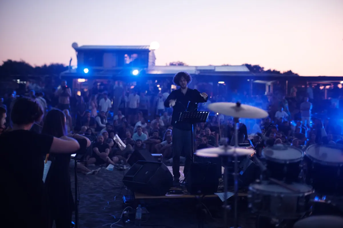 Dirigent podignutih ruku na bini na plaži u sumrak, pred njim publika sjedi na pijesku, u prvom planu bubnjevi i muzičarka