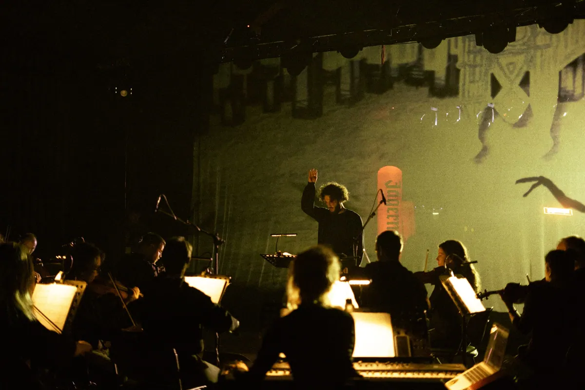 The conductor with a raised arm in front of the orchestra, a projection behind the stage in greenish light