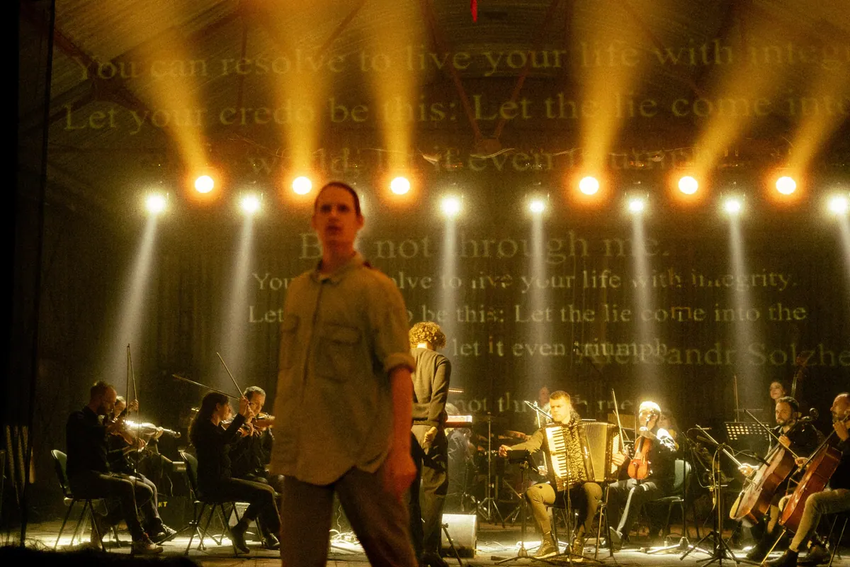 An actor on stage in front of the orchestra, a quotation from Aleksandr Solzhenitsyn projected behind them under spotlights