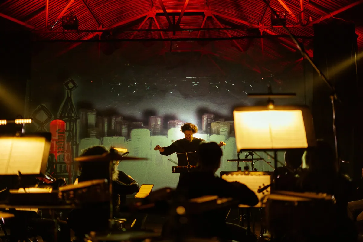 Igor Pejović conducting the orchestra with open arms, a city stage set behind him in red light
