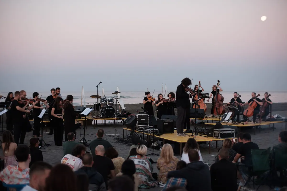 The Montenegro Youth Orchestra playing on an open-air stage by the sea at dawn, the audience sitting on the sand