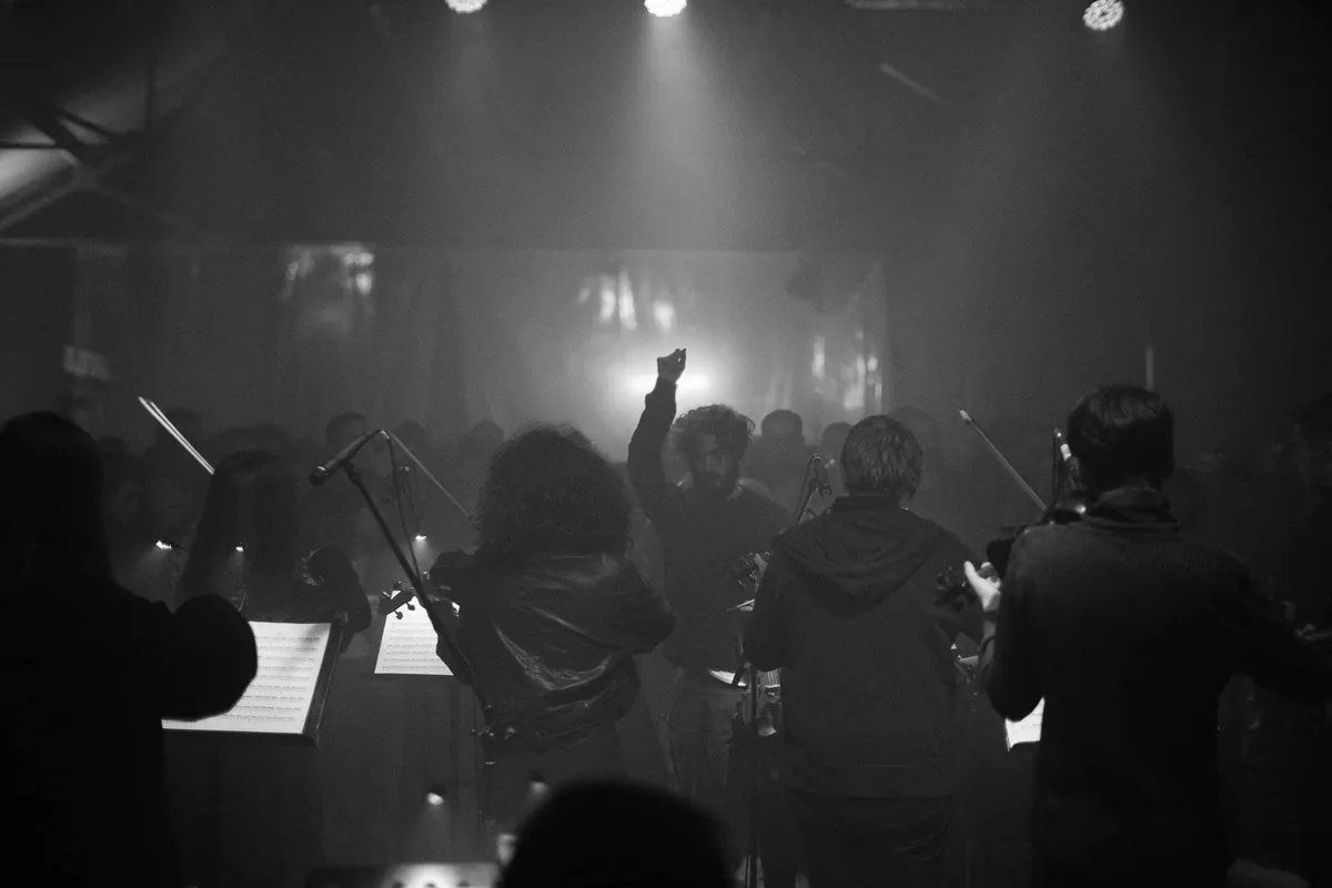 Muzičari sa violinama s leđa pod reflektorima, ispred njih čovjek podignute ruke i publika u izmaglici, crno-bijela fotografija