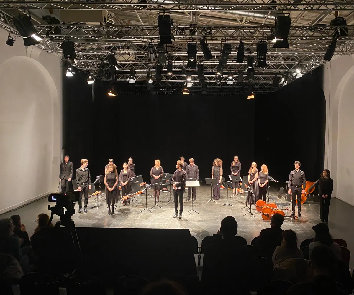 Muzičari orkestra stoje u redu na sceni ispod rasvjetne konstrukcije, publika u mraku u prvom planu