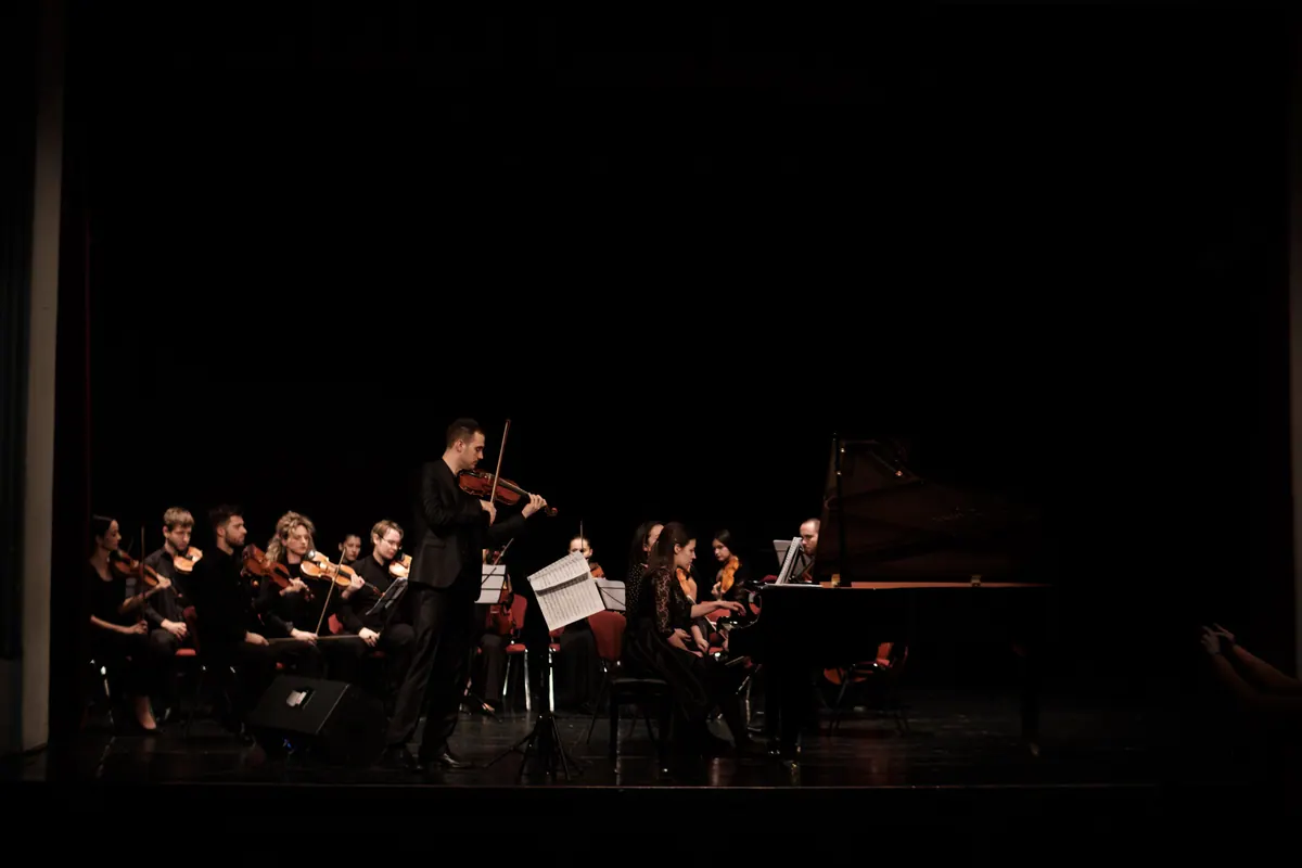 A violin soloist in front of the orchestra, a piano behind